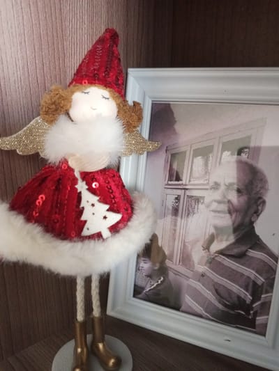 In the foreground of the image, a Christmas-themed angel figurine stands on a wooden surface, possibly a shelf. The figurine has a red, sequined dress with a white trim and a white Christmas tree ornament hanging from its front. It also features golden wings and brown curly hair. To the right of the figurine is a framed photograph. The photograph, which appears to be black and white, depicts an elderly man with gray hair, wearing a striped shirt. Behind him, there is a window with multiple panes. The background is a wooden surface with vertical grain. The image is taken at eye-level.