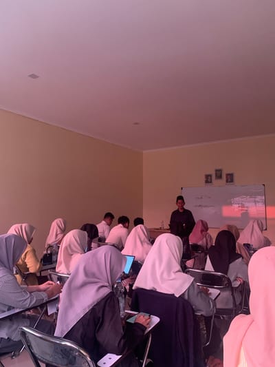 This is a photograph taken indoors, possibly in a classroom or lecture hall, with a warm, pinkish hue cast by the lighting. A man wearing a dark shirt and a peci, a traditional cap, stands at the front of the room, facing a whiteboard. He appears to be a lecturer or speaker. The whiteboard has writing on it, and three framed pictures are hung above it. In front of the speaker, a group of students, predominantly female and wearing headscarves, are seated at desks. Many of them have laptops open, and some are looking at their screens or taking notes. One student in the foreground is looking down at her hands, which are resting on a desk. Another student is holding a phone or small electronic device. The room has cream-colored walls and a white ceiling with recessed square lights. The overall impression is of an educational setting during a lesson. The location is specified as Parung Panjang, Indonesia.