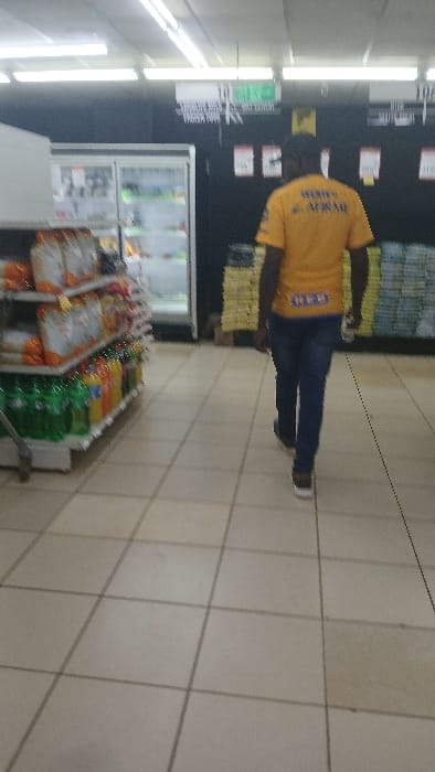 The image depicts the interior of what appears to be a retail store, possibly a grocery or convenience store. The image is taken from a slightly elevated perspective, looking down an aisle.
In the center of the frame, a person is seen from behind, walking away from the camera. They are wearing a bright yellow short-sleeved shirt with blue text printed on the back. While the text is somewhat blurry, parts of it seem to include words like "SERVICE" and "GROUP," with another word that might be "HERMES" or "HERSEY" visible above. The person is also wearing dark blue jeans and light-colored shoes.
To the left of the person (on the left side of the aisle they are walking down), there is a dark-colored wall (likely black or very dark grey). This wall features several white signs or labels, although the text on them is unreadable due to distance and blur. A distinctive green sign with white text is also visible on this wall, but its content is likewise illegible. Further along this wall, a large, glass-doored refrigeration unit is visible, likely containing beverages or other chilled goods. Above the refrigeration unit and along the top of the dark wall, there are shelves with various products, some appearing to be packaged items.
To the right of the person, there is a display aisle featuring multiple racks of products. In the foreground, a display rack holds numerous bottled beverages, with some bottles appearing green and others orange or clear. Behind these bottles, other packaged items, some in orange and white packaging, are visible on shelves.
The floor throughout the store is made of light-colored, square tiles, possibly cream or light grey. The ceiling is equipped with bright fluorescent strip lighting, illuminating the store interior. The overall impression is that of a typical, well-lit retail environment during operating hours. The image is slightly blurry, affecting the readability of smaller details and text.