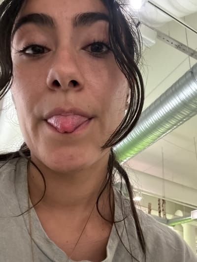 A woman is pictured from the shoulders up, sticking out her tongue. Her dark hair frames her face, and she is wearing a grey t-shirt with a thin gold necklace. The background is out of focus but appears to be an indoor setting with visible ventilation pipes and lighting fixtures. There is no indication of the location in Imatra, Finland.