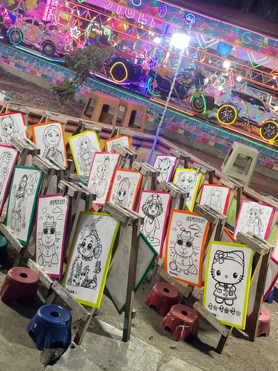 The image depicts an outdoor scene in Kuala Pidie, Indonesia, likely a recreational area or a market at night, given the vibrant neon lights of an amusement ride in the background. The foreground is dominated by a collection of small, framed coloring pages displayed on wooden easels. The coloring pages feature various cartoon characters, including anime-style figures, a teddy bear in a hat and sunglasses, a fish, and Hello Kitty. Each coloring page has a blank line below for a name. Small, colored plastic stools (red and blue) are scattered in front of the easels. The ground appears to be dirt and pavement. The amusement ride in the background is illuminated with colorful lights and shows several miniature vehicles, possibly for children. The overall atmosphere is festive and lively, suggesting a public gathering or event.