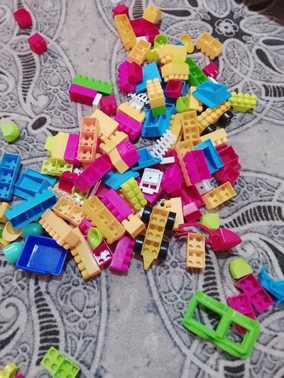 A collection of colorful plastic building blocks is scattered across a gray, patterned surface. The blocks are of various shapes, sizes, and colors, including red, yellow, pink, blue, and green. Some blocks are simple rectangular shapes, while others have more intricate designs with studs and openings. There are also a few wheel-like pieces and a small blue container. The surface appears to be a textile material with an intricate, possibly floral or paisley, pattern. The lighting is overhead, casting soft shadows. There are no people or animals visible in the image. The image captures a close-up, overhead view of the toy blocks. The scene does not provide any information about the location in the city of Kümüş-Aziz, Kyrgyzstan.