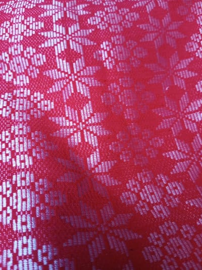 The image depicts a red fabric with a woven floral pattern. The pattern consists of repeating star-shaped motifs formed by small, rectangular blocks that give the fabric a textured look. The "stars" have a subtle sheen, contrasting with the matte red background. The fabric appears to be draped, with light reflections creating variations in color intensity. The overall impression is that of a decorative, perhaps traditional textile.