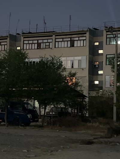 The image depicts a multi-story apartment building at dusk in Şamaldı-Say, Kyrgyzstan. The building has a light-colored facade with multiple windows, some of which are illuminated. A dark-colored pickup truck and a minivan are parked in front of the building, partially obscured by trees. The ground in front of the vehicles appears to be unpaved, with scattered debris. In the background, other buildings and trees are visible, suggesting a residential area. The sky is a deep blue, indicating the transition from day to night. There are no people visible in the image.