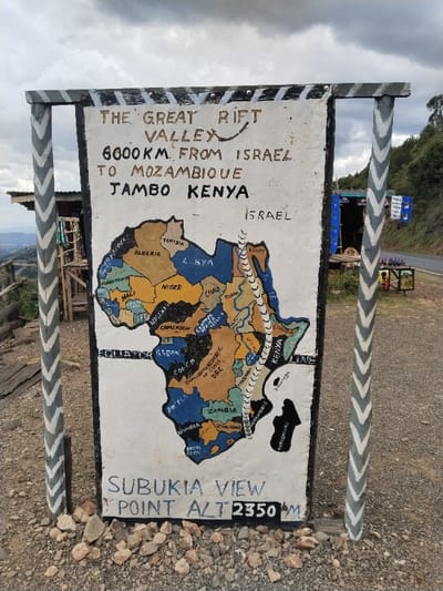 The image displays a weathered sign board marking "SUBUKIA VIEW POINT ALT 2350 M" in Waseges ward, Kenya. The board, supported by two wooden posts with grey and white chevron patterns, features a white background. The upper section states: "THE GREAT RIFT VALLEY 6000KM. FROM ISRAEL TO MOZAMBIQUE JAMBO KENYA", with "ISRAEL" indicated separately to the right. Below this, a hand-painted map of Africa is depicted, with various countries colored differently in tan, blue, light green, and dark blue. A prominent white, wavy dashed line, representing the Great Rift Valley, extends from the area of Israel (off the map's top edge) southward through eastern African countries, including Kenya and Tanzania, towards Mozambique. The "EQUATOR LINE" is marked across the map.
The sign is situated on loose gravel and small rocks. In the background to the left, a rustic wooden structure with a corrugated metal roof is visible, overlooking a distant valley. To the right, a paved road runs horizontally, beyond which are green, tree-covered hillsides. Near the road, a blue and white stall or vendor structure is discernible. The sky above is overcast with grey and white clouds. No individuals are present in the scene.