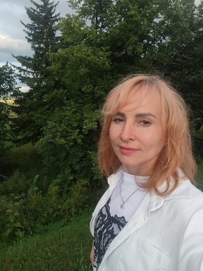 A medium shot captured from a selfie angle shows a Caucasian woman with blonde, shoulder-length hair and light skin. She is wearing a white collared shirt over a white t-shirt with a black lace pattern and a silver necklace with a pendant. She has light makeup on and is looking slightly to her left. The background is a lush green landscape with tall trees, likely in the city of Staraya Yum'ya, Russia. The lighting suggests it is daytime, possibly late afternoon due to the soft light.