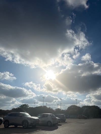 The image captures a parking lot in Beit Yizhaq - Shaar Hefer, Israel, under a dramatic sky. The foreground shows a paved surface with several parked cars, including a silver SUV prominently positioned on the left, and a smaller silver hatchback to its right. In the background, a line of trees forms a dense canopy, with tall poles supporting stadium lights visible above them. The sky is a dynamic mix of bright sunlight breaking through scattered clouds, casting rays across the scene and illuminating parts of the landscape. The overall impression is a partly cloudy day with intense sunlight.