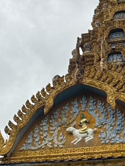 The image shows a close-up view of the ornate golden roof of a temple, likely in Sneung, Cambodia. The roof features intricate carvings and decorative elements, typical of Khmer architecture. Prominently displayed is a relief sculpture depicting a green-skinned figure riding a white rabbit, surrounded by stylized floral or cloud-like patterns in a pale blue color. The overall color palette is dominated by gold, with accents of blue and green. The sky above is overcast and appears white or light gray. The architecture suggests a place of religious or cultural significance.