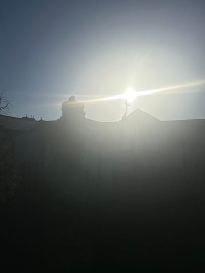 This is a vertically oriented, overexposed photograph taken during daytime, likely in the late afternoon due to the low angle of the sun. The scene features the sun prominently in the upper left quadrant, emitting intense rays that create a strong lens flare and wash out much of the image with bright light. Silhouetted against the glare are parts of buildings and trees, suggesting an outdoor, possibly urban or suburban, setting.
In the foreground, the tiled roof of a building is visible, along with several chimneys. Further back, other structures appear, though their details are obscured by the sunlight and shadow. A tree or dense foliage is also present, partly silhouetted and illuminated by the sun. The overall impression is one of a bright, possibly hazy day. The picture appears to be taken from a low vantage point, looking upwards. No people are visible in the image, and there is no visible text. The location context provided indicates Środa Wielkopolska, Poland.