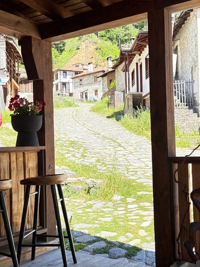 The image shows a view from a wooden veranda in the village of Shiroka Laka, Bulgaria. In the foreground, two wooden bar stools are visible on a wooden deck. To the left, a dark planter filled with red flowers sits on a wooden shelf. Beyond the veranda, a cobblestone street winds uphill, lined with traditional stone houses. Green vegetation, including grass and trees, grows alongside the street and on the surrounding hillsides. The architecture suggests a historic Balkan village setting.