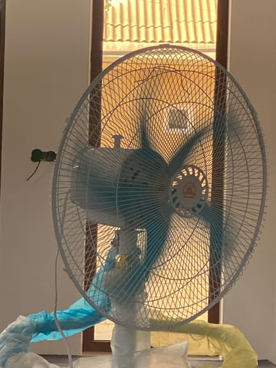 The image shows a white electric fan with blue blades, positioned in front of a window with orange blinds. The fan is partially covered with blue and yellow plastic sheeting, suggesting it is either being protected during construction or cleaning, or is being stored. A small green electrical outlet with a wire is visible on the wall to the left of the fan. The overall lighting suggests it is daytime, with sunlight filtering through the blinds. The scene appears to be indoors, likely in a residential or commercial building, but no specific location in Arbagish, Uzbekistan, can be identified from the image itself.