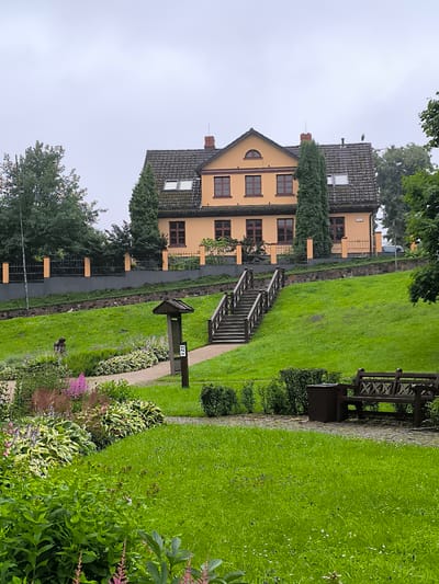 The image depicts a residential building with an orange facade, a dark-tiled roof, and multiple windows, situated on an elevated grassy slope. A prominent wooden staircase with railings ascends the slope towards the building. In the foreground, a well-maintained garden features various plants, including flowering bushes and a paved walkway. A wooden bench and a trash receptacle are visible on the walkway. The sky is overcast. This location is the Kuldīga District Museum in Kuldīga, Latvia, also known as the Kuldīga Local History Museum.