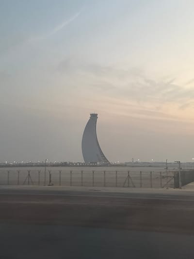 The image depicts an expansive airport landscape in Al Matar, United Arab Emirates, under a hazy sky exhibiting a gradient from pale blue-gray overhead to a light peachy tone near the horizon. The dominant subject is a tall, white air traffic control tower with a distinctive curved, sail-like or crescent-shaped design, culminating in an observation cabin and an antenna array at its summit. In the immediate foreground, a dark, paved surface occupies the bottom quarter of the frame. Beyond this pavement, a flat, light brown, sandy terrain extends horizontally, segmented by a multi-strand security fence composed of vertical posts and mesh panels, with some sections featuring angled stabilizers. Along the distant perimeter of the airport, numerous small, white lights are visible. The scene is devoid of people or discernible actions.
