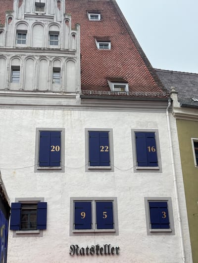 The image displays the exterior of a multi-story building facade in Meissen, Germany, under an overcast sky. The building's lower and mid-sections feature a textured white stucco finish. On the upper floor, three windows are visible. From left to right, these windows are fitted with closed dark blue shutters bearing gold numbers: "20" on the left, "22" in the center, and "16" on the right. On the floor below, three additional windows are present. The leftmost window has partially open dark blue shutters, revealing a dark interior. The central window features closed dark blue shutters, with a gold "9" on the left shutter and a gold "5" on the right shutter. The rightmost window on this level has closed dark blue shutters, with a gold "3" visible on the right shutter. Directly below the central lower window, a dark-colored sign with the word "Ratsteller" in an ornate, old German script is mounted on the facade. Above the white stucco section, an ornate white stone or stucco gable with multiple arched recesses and small, rectangular windows extends upwards. To the right of this gable, a steeply pitched roof covered in red tiles is visible, featuring several small dormer windows. A portion of an adjacent building, with a lighter-colored wall and a dark tiled roof, is visible on the far right, separated by a white vertical downspout. The scene contains no individuals or active elements.