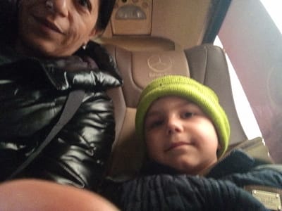 The image captures two individuals, an adult female and a child, seated within a vehicle cabin. The adult female, with dark hair, occupies the left side of the frame, visible from the chest up. She wears a black, glossy puffer jacket and a grey seatbelt across her chest. Her right index finger is partially visible in the bottom foreground, suggesting she is holding the camera. To her right, a child, appearing to be between 5 and 8 years old, is seated. The child wears a bright lime green knit beanie and a dark puffer jacket, looking directly at the camera with a neutral expression. Behind the child, a light-colored vehicle seat with an embossed Mercedes-Benz logo on the headrest is visible. An overhead panel and a light-colored window or interior panel are visible in the background on the right. The precise location within the city of Babuk, Bulgaria, cannot be determined from the visual information presented in this image.