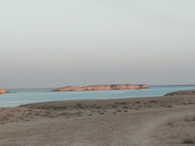 The image depicts a serene, natural landscape with a body of water and barren, sandy terrain. In the foreground, a dry, dusty path leads into a desert-like environment sparsely populated with small, dry bushes. The middle ground features a calm, light blue body of water, likely a lake or reservoir. Across the water, a prominent, elongated island or peninsula rises, characterized by its reddish-brown, layered rock formations, suggesting a geological composition of sedimentary rock or dried mud. The island appears rugged and uninhabited. The background consists of a hazy, pale sky, indicative of early morning or late afternoon light, with faint, distant structures visible on the horizon to the far right, hinting at human presence beyond the immediate natural setting. The overall impression is one of a vast, arid environment with a stark contrast between the dry land and the tranquil water.