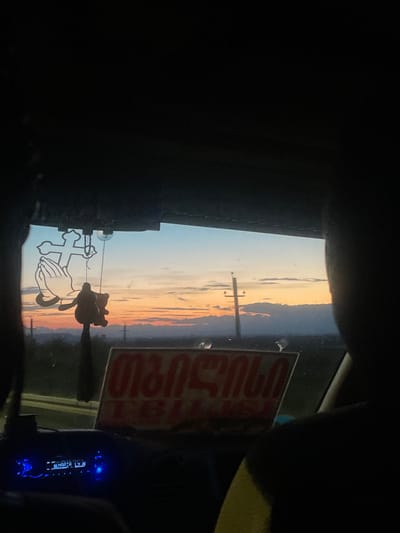 The image captures a view from inside a vehicle, looking out the windshield at a sunset. The sky displays a gradient of colors, from a pale blue at the top to hues of orange and pink near the horizon, with scattered clouds. In the foreground, hanging from the rearview mirror, is a religious decal featuring a cross and praying hands, along with a small, brown teddy bear charm. A red and white sign with Georgian text is visible on the dashboard. The text on the sign appears to be "თბილისი", which translates to Tbilisi, indicating a possible destination or origin. The radio display shows "MARIA LY" in blue illuminated text. The overall scene suggests a travel moment at dusk, possibly in or near Akhalsheni, Georgia, given the sign.
