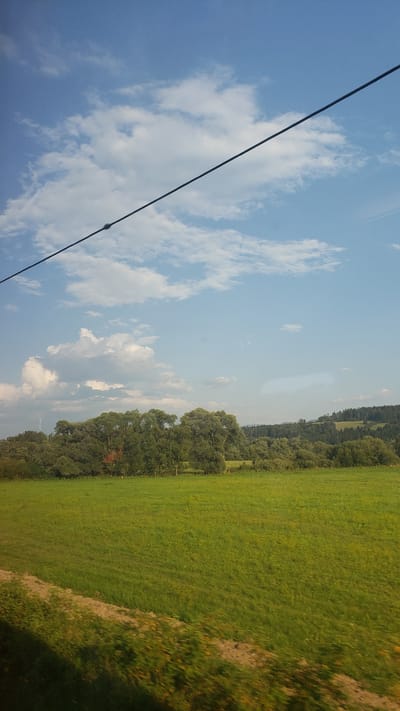 The image depicts a vast, green meadow in the city of Turík, Slovakia, under a bright blue sky dotted with white clouds. A thin black wire stretches diagonally across the upper portion of the frame. In the distance, a dense line of trees forms a natural border along the horizon, with rolling hills covered in darker foliage extending beyond them. The foreground shows a blurred section of the meadow, suggesting movement, possibly from a vehicle.