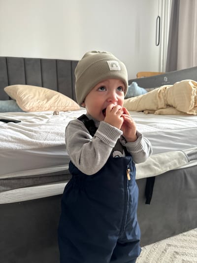 A young child stands in an indoor bedroom environment. The child wears a light olive ribbed beanie with a rainbow patch, a grey long-sleeved sweater, and dark blue overalls. Both hands are raised to the mouth, obscuring the chin, and a small, dark object is partially visible within the mouth. The child's gaze is directed upwards and to the right.

The background features a bed with a dark grey upholstered headboard composed of vertical panels. The bed is covered with a white textured mattress protector or fitted sheet. A beige pillow rests on the left side of the bed, a light blue object resembling a pillow or plush toy is near the center, and a crumpled beige duvet or comforter is on the right side. A black remote control lies on the bed to the left of the child, and a small, light-colored decorative object, possibly an animal figurine, is located further back on the bed. A light-colored carpet with a subtle pattern covers the floor. The walls are light-colored. To the right, a grey curtain or blind is partially visible, and a black strap or cord hangs vertically. A light-colored mesh bed rail or barrier is positioned adjacent to the bed on the right, above a dark grey storage component or bed frame base.