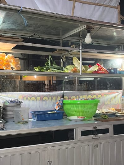 The image shows the interior of a food stall in Denasri Kulon, Indonesia. The stall is equipped with shelves containing fresh produce, including green onions, cabbage, and what appear to be oranges or tangerines packaged in plastic. There are also stacks of plates, a blue basket, and a green colander on a counter. The lighting suggests it is nighttime or indoors. The background displays a blurred view of outdoor elements, possibly a street or another building, with lights reflected on a wet surface, indicating recent rain. A prominent banner with yellow lettering is visible behind the counter. The stall appears to be a commercial setup for selling food.