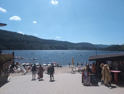 The image shows a sunny day scene at the Titisee lake in Titisee-Neustadt, Germany. The foreground features a paved walkway with people strolling and sitting at tables. To the right, there is a small building, possibly a cafe or kiosk, with a humorous hot dog mascot standing outside. A menu board lists items like "Softeis," "Chicken Nuggets," and "Cocktails."
The middle ground depicts a sandy beach area with lounge chairs and people relaxing. Numerous small boats and paddle boats are visible on the shimmering blue water of the Titisee. In the background, lush green hills covered in trees form the backdrop to the lake, under a clear blue sky with a few scattered clouds. The overall atmosphere is one of leisure and outdoor recreation.