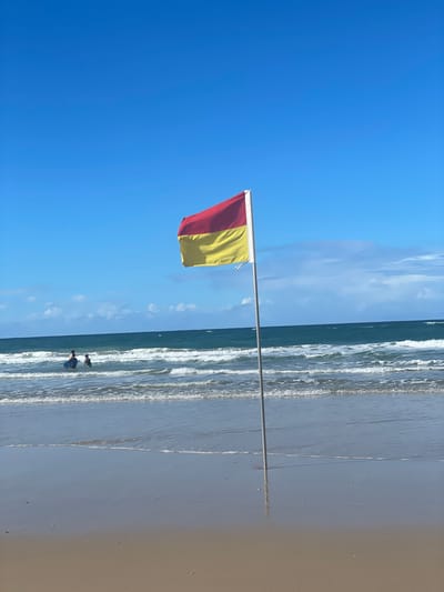 A red and yellow flag flies on a metal pole in the foreground of a beach scene in Surfers Paradise, Australia. The flag is positioned to the right of the frame, with its colors, red on top and yellow on the bottom, clearly visible against a bright blue sky with scattered white clouds. In the middle ground, the ocean stretches towards the horizon, with waves breaking on the shore, creating white foam. Two figures, appearing to be children, are wading in the shallow water, their bodies partially submerged. The beach itself is composed of light-colored sand, wet near the water's edge, reflecting the sky. The overall atmosphere is sunny and inviting, suggesting a pleasant day at the beach.