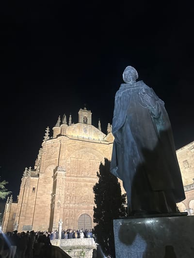 The image displays a nighttime scene in the Plaza de San Esteban, Salamanca, Spain. In the foreground, positioned to the right, is a large bronze statue of Fray Luis de León. The robed figure stands with his head slightly tilted left and upward, appearing to hold a book or scroll in his left hand. The statue rests on a dark stone pedestal, partially visible, with a faint shadow cast on it.

Dominating the midground and left background is the illuminated facade of the Convento de San Esteban. This large, historic sandstone building features intricate Baroque and Plateresque architectural detailing, including a grand arched entrance, multiple tiers of decorative relief carvings, and a central dome crowned with a lantern and bell tower. The structure is brightly lit with warm artificial light, contrasting against the dark, clear night sky.

Below the illuminated convent facade, a dense crowd of approximately 50 to 70 individuals is gathered on a raised platform or steps. The people appear as indistinct figures, predominantly facing towards the convent's entrance. A dark, slender conifer tree stands between the statue and the building, with additional sparse foliage visible on the far left. The scene captures an evening gathering in a historic urban environment.