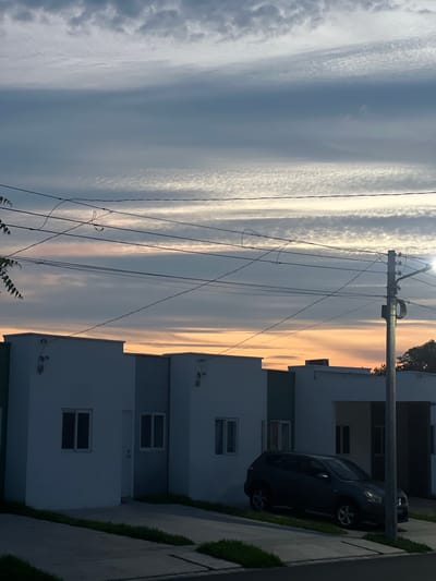 The image depicts a suburban street scene in San Miguel, El Salvador, during dusk. The sky is filled with a dramatic display of clouds, characterized by horizontal bands of varying shades of grey, white, and a soft orange-pink hue near the horizon, suggesting the sun is setting. A complex network of power lines crisscrosses the upper portion of the frame, suspended from a utility pole visible on the right side.
In the foreground, a row of modern, minimalist houses with white facades and green accents line the street. The houses appear to be identical or very similar in design, with rectangular windows and flat roofs. A dark-colored SUV is parked in the driveway of one of the houses. The ground is paved with concrete, with narrow strips of grass separating the pavement from the houses. The overall impression is one of a newly developed residential area, quiet and orderly. There are no people visible in the image.