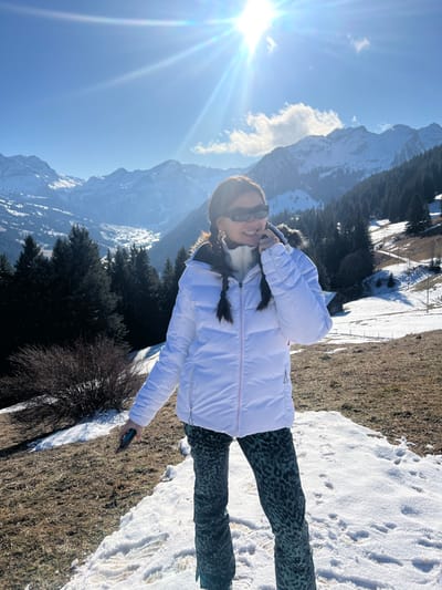 This image captures a female individual standing on a partially snow-covered, grassy hillside in the mountainous region of Saanen, Switzerland. The scene is illuminated by a bright sun, visible as a prominent sunburst in the upper right quadrant of the clear blue sky, which contains a few white clouds.
The subject, of apparent East Asian descent, is positioned slightly right of center, facing forward with a slight turn of her head to the left. She wears a white puffer jacket with a visible zipper closure and a brown fur-trimmed hood, a white high-collared garment underneath, and dark green trousers featuring a leopard or camouflage pattern. Her dark hair is styled into two braided pigtails that fall over her shoulders. She wears dark, rectangular sunglasses and displays a smiling facial expression. Her right hand is raised, with the index finger touching her lower lip, while her left hand holds a dark-colored smartphone, pointed downwards at her side.
The foreground consists of patches of brown grass interspersed with snow, exhibiting visible footprints. The background features a deep valley surrounded by rugged, snow-capped mountains and dense coniferous forests. A small, dark wooden structure is discernible on a distant, snow-dusted slope in the mid-right background.
