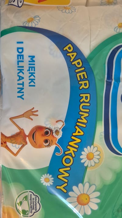 The image shows a close-up of a plastic-wrapped package of chamomile toilet paper, likely manufactured in Poland. The packaging features a cartoon ant character wearing a flower on its head, waving with one hand. The ant is orange with large, expressive eyes and is smiling.

The text on the packaging is in Polish. Prominently displayed in large yellow letters with a blue outline is "PAPIER RUMIANKOWY," which translates to "Chamomile Paper." Below this, in smaller blue letters, are the words "MIĘKKI" and "I DELIKATNY," meaning "Soft" and "Delicate," respectively.

The background of the packaging is a light blue sky with stylized white daisies, some with yellow centers. A green wavy border runs across the top and down the sides. In the lower right corner, there is a circular green emblem with text that appears to relate to environmental commitments, possibly "Zobowiązanie do Zerowej Emisji CO₂ NETTO," indicating a commitment to net-zero CO₂ emissions.

The overall impression is one of a soft, gentle, and perhaps eco-conscious product, marketed with a friendly and whimsical character. The lighting suggests an indoor setting, possibly a store or home.