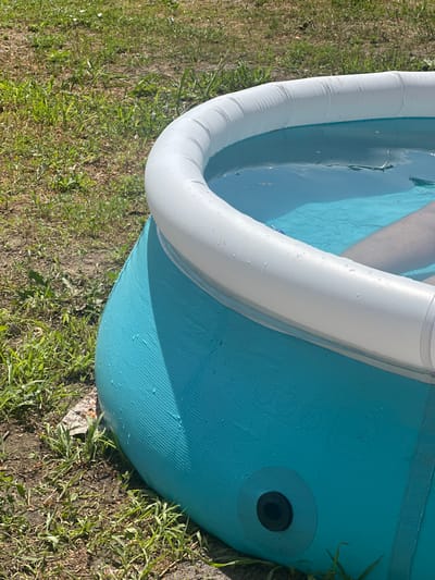 The image shows a close-up, oblique view of a blue inflatable pool filled with water, situated outdoors on a patch of grass. The pool has a white inflatable top ring. A person's legs are visible submerged in the water, indicating they are in the pool. The ground around the pool is covered with dry grass and dirt, with some green vegetation. The setting appears to be a residential area, likely a backyard. The pool has a black drain plug visible on its side. The lighting suggests it is daytime, with sunlight casting shadows. The location is identified as Yablunivka, Ukraine.