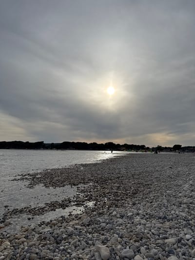 The image captures a serene coastal scene in Općina Medulin, Croatia. The sky is overcast with the sun breaking through, casting light across the water and pebble beach. A line of vegetation runs along the horizon, separating the beach from what seems to be a built-up area. The beach is composed of numerous pebbles and is partially submerged in water.