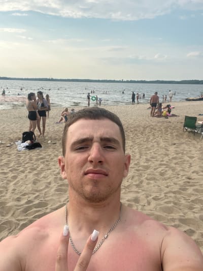This is a selfie taken at a beach on a bright day, likely in the afternoon, judging by the lighting. The foreground is dominated by the upper body of a young man, facing the camera. He has short brown hair and is wearing a silver chain necklace. His skin appears tanned and slightly sunburnt on his shoulder and chest. Beside him, a hand with white nail polish is giving a peace sign.
The background depicts a crowded beach scene. Numerous people are visible, some wading in the shallow water, while others are relaxing on the sand. The water is calm and appears to be a lake or a large river. There are several beach chairs and umbrellas scattered across the sand, indicating a popular recreational area. The overall atmosphere seems relaxed and summery, with people enjoying the warm weather. The terrain is sandy, typical of a beach. No text is visible in the image.