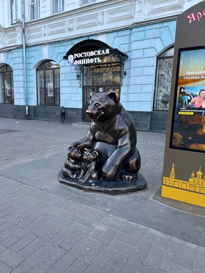 The image shows a bronze sculpture of a mother bear with two cubs, located in front of a building with light blue and white painted facade. Above the entrance of the building, there is a black awning with white Cyrillic text that reads "РОСТОВСКАЯ ФИНИФТЬ". To the right of the sculpture and building, a digital billboard displays an advertisement. The billboard shows a woman participating in a race and text in Russian. The ground in front of the sculpture and building is paved with grey cobblestones. The scene appears to be a street or pedestrian area in a city.