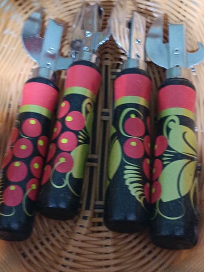 This is a close-up, eye-level, indoor photograph of four vintage can openers. The can openers have red and black handles decorated with a folk art pattern of red circles with yellow dots, green swirls, and green leafy shapes. The metal parts of the can openers are silver and are visible at the top of the image. The can openers are resting in a woven basket, likely made of wicker, which occupies the majority of the background. The lighting is dim and casts soft shadows. There are no people visible in the image. The image does not provide any information about the location in the city of Martuni, Armenia.