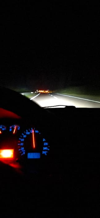 This image depicts a nighttime scene from the driver's perspective inside a vehicle traveling on a road in Majmaa Tolba, Morocco. The foreground shows a partial view of the vehicle's dashboard, with the speedometer prominently illuminated in blue and red. The speedometer needle indicates a speed of approximately 110-115 km/h. A red warning or indicator light is illuminated on the far left of the dashboard. The steering wheel rim is partially visible on the left. The midground displays a dark asphalt road illuminated by the vehicle's headlights, revealing white lane markings on both sides. The road appears straight and clear. In the distant background, several clusters of orange and red lights are visible, suggesting the presence of other vehicles ahead or distant infrastructure. The environment outside the immediate headlight beam is dark, indicating a lack of ambient light.