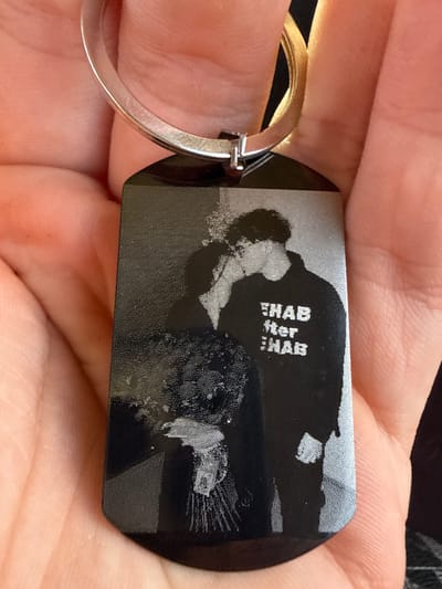 The image shows a close-up of a black, rectangular dog tag keychain held in a person's hand. The keychain has an engraved image of what appears to be two people embracing. The engraving is in black and white, creating a stark contrast against the black background of the tag. Some of the engraving is textured, with a speckled appearance, while other parts are smoother.
There is visible text engraved on the tag, arranged vertically. The text reads "REHAB after REHAB". The keychain is attached to a silver ring. The background of the image consists of the person's skin, showing fingerprints and the texture of their hand, as well as the dark fabric of clothing. The lighting suggests an indoor setting, with no indication of time of day or weather. The overall mood conveyed is personal, perhaps sentimental, given the nature of the engraved image and text.