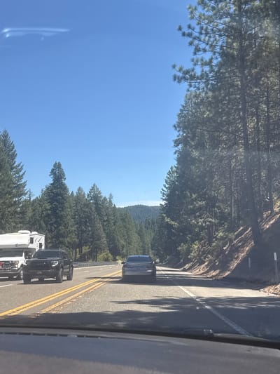 A vehicle is traveling on a highway lined with tall pine trees. The highway has two lanes in each direction, marked with yellow lines. On the left side of the road, a white RV is parked alongside a black Toyota Tacoma truck. The sky is clear blue with a faint white cloud formation visible. The terrain slopes upwards on the right side of the highway, covered in trees and vegetation. The image appears to be taken from the perspective of another vehicle on the highway. This scene is characteristic of Highway 50 near Pollock Pines, California, a mountainous area with dense forests.