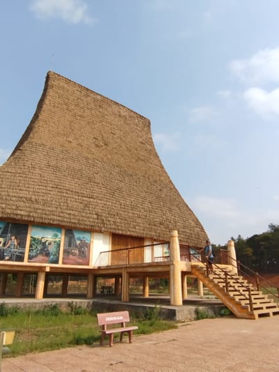 The image depicts a traditional, stilt-supported building with a high, steeply pitched, thatched roof. The building is adorned with large murals. A man stands on the staircase leading to the building's entrance. The structure is surrounded by landscaped grounds with a bench and paved areas. The sky is clear with a few clouds. The location is inferred to be in Đà Giang, Vietnam, based on the building's style.