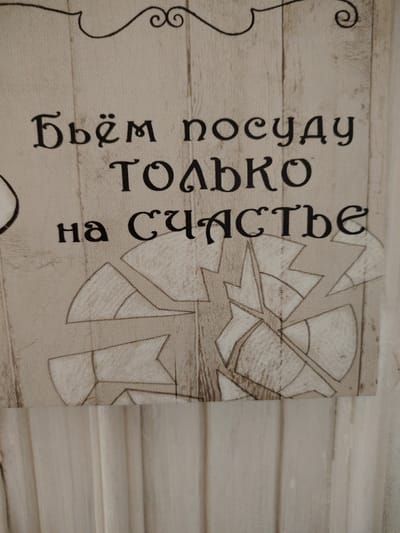 The image shows a close-up of a textured surface with Cyrillic text printed in black. The text appears to be a decorative element, possibly on a wall hanging or fabric. The words read "Бьём посуду только на счастье" which translates to "We break dishes only for good luck." Below the text, there is a stylized graphic that resembles broken pieces of pottery or glass arranged in a circular pattern. The background has a wood-grain texture. The image does not contain any elements related to the city of Vladimirovka, Russia.