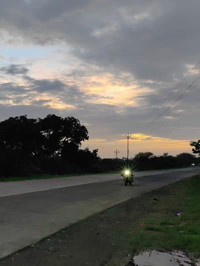 An overcast sky with patches of light illuminates a rural road in Dattapur, India. A motorcycle with its headlight on, emitting a bright starburst effect, travels down the asphalt road. Silhouetted trees and vegetation line the left side of the road, while a grassy verge with a puddle of water is visible on the right. Utility poles and wires stretch across the background, contributing to the atmospheric scene.