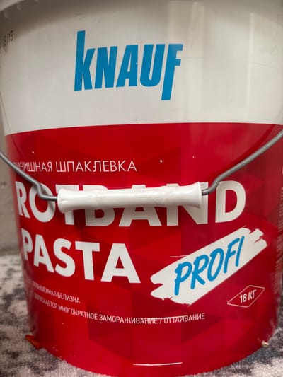 A close-up, eye-level shot shows a white plastic bucket with a red label. The brand name "KNAUF" is emblazoned in large, blue capital letters across the top of the bucket. Below this, in smaller white lettering on the red label, it reads "ФИНИШНАЯ ШПАКЛЕВКА" which translates to "FINISHING PUTTY". Dominating the center of the label are the words "ROTBAND PASTA" in large white capital letters, with "PROFI" written in blue cursive script on a white, paint-splatter-like background to the right. A silver metal handle with a white plastic grip is attached to the top of the bucket. Towards the bottom right, a red diamond shape encloses the text "18 КГ", indicating the weight. Russian text runs along the bottom of the label, including "ПОВЫШЕННАЯ БЕЛИЗНА" ("INCREASED WHITENESS") and "ДОПУСКАЕТСЯ МНОГОКРАТНОЕ ЗАМОРАЖИВАНИЕ / ОТТАИВАНИЕ" ("MULTIPLE FREEZING / THAWING IS PERMITTED"). The bucket rests on a grey and white patterned surface, possibly a rug or carpet. The background is a plain, light-colored wall, with a faint shadow line running horizontally across it. The lighting is bright and even, suggesting an indoor setting, possibly a store or storage area in Posyolok Imeni Morozova, Russia. There are no people or animals visible in the image.