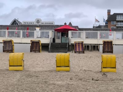 This is a wide, eye-level, outdoor, daytime shot of a beach in Bergen, Netherlands. The sand is a light brown color and appears to be flat and even, with a small pile of sand in the foreground. In the center of the frame, there are three yellow wicker beach chairs with their hoods up. In the background, there is a building with a wooden facade and large windows, housing a restaurant or cafe. A bright red umbrella is open in front of the building. Several striped beach chairs are visible between the building and the yellow chairs. Two flags are flying on poles above the building. The sky is overcast and gray.