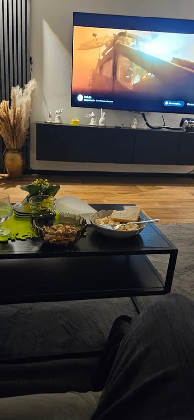 The image shows a living room setting in Gütersloh, Germany. A television displaying a dynamic orange and white scene hangs on the wall above a black, horizontally-slatted base. A low, black shelf unit with decorative ceramic bunnies extends along the wall. A black table sits nearby with a bowl of bread and salad, a container of nuts, and a small vase of yellow flowers on a bright green doily. The scene is well-lit and cozy. A sofa is partially visible on the right. There are no people visible in the scene.