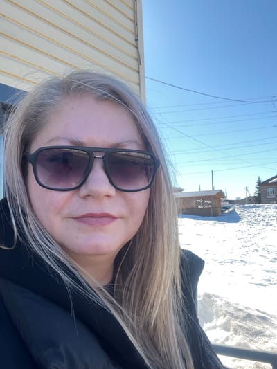 A female individual is depicted in a close-up selfie in Сундур, Russia, under a clear blue sky. She has long, light blonde/greyish hair and wears dark, rectangular sunglasses with thick black frames. Her expression is neutral, and she has light pink lipstick applied. She is wearing a black, insulated jacket. To the immediate left, a building with light-colored horizontal siding is partially visible. The background shows a snow-covered landscape indicative of winter. In the mid-ground, a wooden gazebo with a dark, sloped roof and lattice sides is present. Further to the right, a multi-story wooden house with a visible window stands on a slight incline, partially obscured by snow. Multiple utility poles and overhead wires traverse the scene against the clear blue sky.