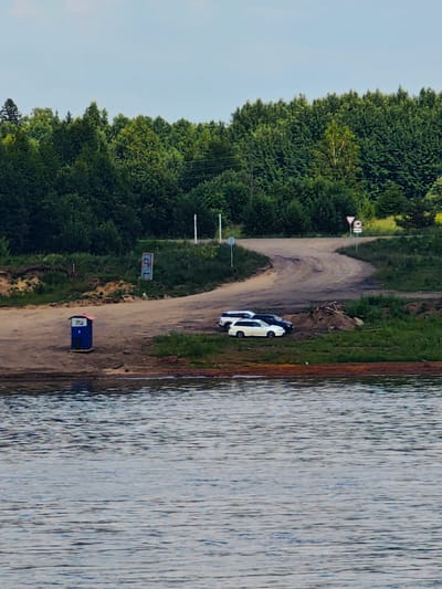 A body of water occupies the foreground of the image, with a brown shore beyond. A small blue structure with a red roof sits on the shore. Further inland, a dirt road curves uphill, passing several parked cars. The road is flanked by green grass and trees. In the background, a dense forest stretches across the horizon. The sky is a pale blue. The location is the Erkalovo, Russia area.