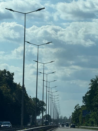The image depicts a view from a vehicle, likely in motion, traveling along a highway. The perspective is slightly angled, showing a long row of streetlights receding into the distance. The sky is filled with dramatic, large cumulus clouds, suggesting a partly cloudy or overcast day. Lush green trees line the right side of the road, with more trees visible in the background, interspersed with some buildings. Several cars are also visible on the highway in the distance, suggesting moderate traffic. The scene appears to be set during daylight hours.
There are no people clearly visible. The primary objects are the streetlights, the road, trees, and vehicles. The setting is a highway in a semi-rural or suburban area, characterized by the presence of both infrastructure (road, streetlights) and natural elements (trees, sky).
The activity taking place is travel along a highway. There are no explicit emotional cues. The weather appears to be dynamic due to the prominent cloud formations. The location is identified as Vinnytski Khutory, Ukraine, though this context is external to the visual information in the image itself. A blue sign with text is partially visible on the right side, but the text is illegible in the provided image.