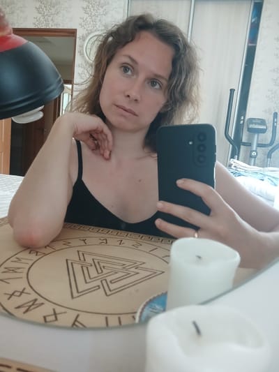 A young woman with curly brown hair and green eyes takes a mirror selfie in a room. She is wearing a black tank top and has her right hand resting on her chin. Her left arm is bent at the elbow and rests on a wooden board with runes and a symbol that resembles a Valknut. In front of the board are two white candles, one in focus and the other blurred. Behind her, a mirror reflects an exercise machine and a closet with sliding doors. The background shows patterned wallpaper. The environment appears to be a bedroom in a residential setting. The woman is indoors, likely in her home in Maslova Pristan', Russia.