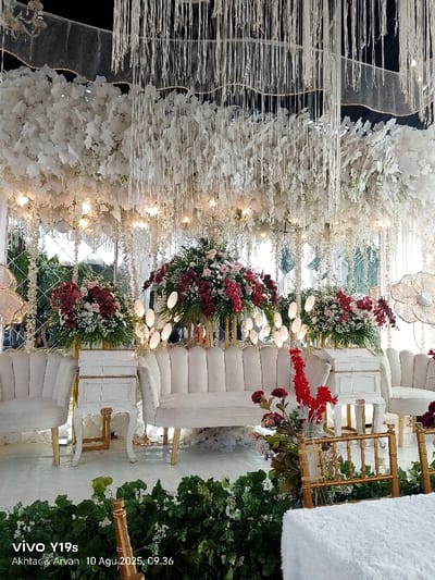 The image displays an elaborately decorated indoor or semi-outdoor event space, located in Jagapura Kulon, Indonesia, captured on August 10, 2025, at 09:36 AM.
The scene is dominated by a central seating arrangement, likely a bridal dais or pelaminan, positioned against a reflective backdrop composed of square or diamond-shaped mirrored panels. Overhead, a dark ceiling is obscured by a dense canopy of white artificial foliage, from which numerous thin, white, vertical strips or ribbons descend. Multiple small, warm-toned lights are interspersed within the white foliage, providing illumination.
The main seating area features a cream-colored, upholstered two-seater sofa with a fluted back, flanked by two matching single armchairs. Between the sofa and each armchair stands a white, ornate side table or pedestal with gold accents. Behind these tables, two large floral arrangements are displayed on tall, gold-colored stands. These arrangements are densely packed with deep red/maroon, pink, and white flowers, accented by green foliage. Numerous small, round, white light fixtures are integrated into the decor around these floral arrangements.
The lower foreground shows a border of artificial green foliage interspersed with small white flowers. A gold-colored, bamboo-style chair frame and a portion of a white, textured object (possibly another seat or cushion) are visible in the lower right. No individuals, direct actions, or interactions are visible within the frame; the scene appears set and static.