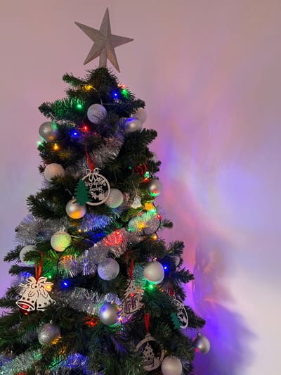 The image displays a decorated artificial Christmas tree positioned indoors in Iława, Poland. The tree is dark green and adorned with a silver, glitter-covered five-pointed star topper. A string of multi-colored lights (red, green, blue, yellow) is wrapped around the branches. Silver metallic tinsel garlands are draped throughout the tree. Various ornaments are present, including numerous spherical balls in silver, grey, and white, some smooth and reflective, others textured. Several wooden cutout ornaments are visible: one prominent circular ornament with "Wesołych Świąt" (Merry Christmas) text and a green glitter tree, a double bell design with cutouts, and other stylized Christmas tree shapes, some with internal scene details, and a small angel figure. These ornaments are suspended by red ribbons or clear string. The tree stands against a plain, light-colored wall which exhibits soft, vertical streaks of colored light (purple, blue, red, yellow) reflecting from the tree's lights. No individuals, animals, or dynamic actions are depicted. The scene is static, focused solely on the decorated Christmas tree and its immediate background.